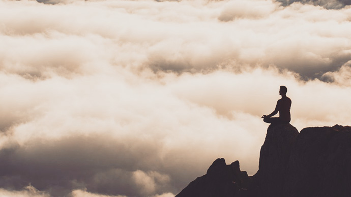 Man in yoga lotus position on rocky outcropping, clouds in the background