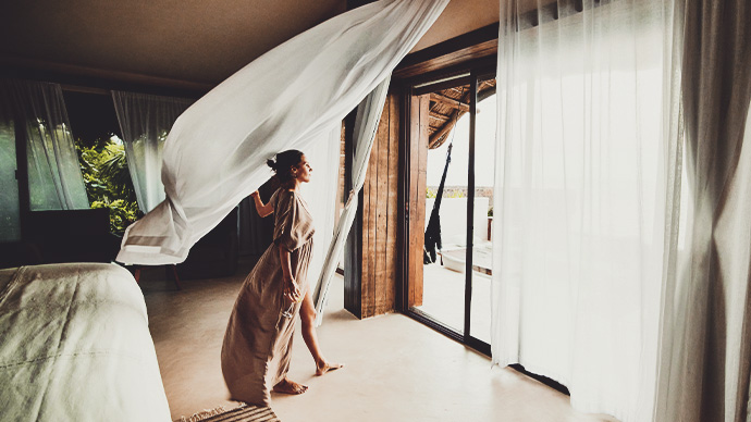 Wide shot of woman standing in luxury hotel suite looking at view with curtains blowing in wind