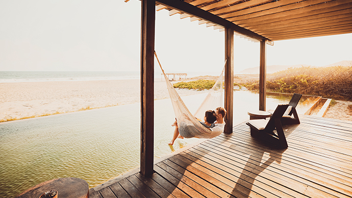 Extreme wide shot of romantic couple relaxing in hammock beside pool at luxury beachfront hotel overlooking ocean while on tropical vacation