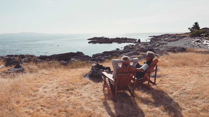 Couple relax on wooden chair, look out to calm sea-At sunrise, bay and coastline distant