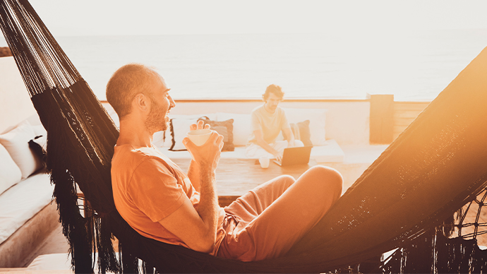 Medium wide shot of man sitting in hammock watching sunrise while partner works on laptop from deck of luxury tropical beach villa