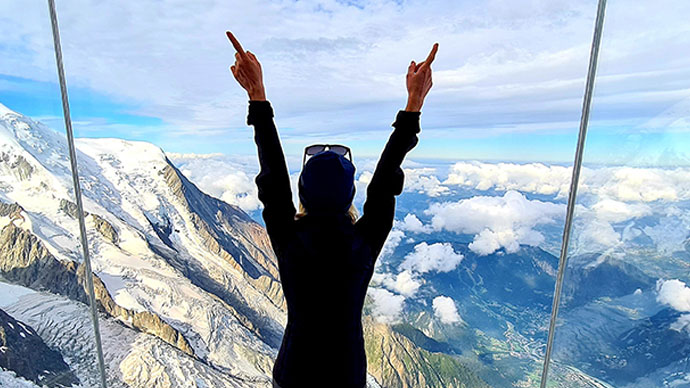 Back of woman looking out window on top of snow covered mountains, both hands doing the #1 sign 