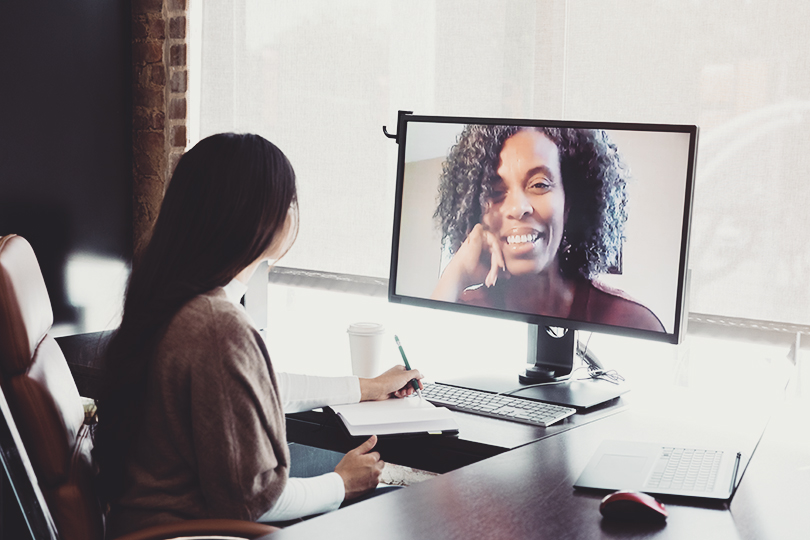A young businesswoman talks via video call with a mature female colleague. The colleague is telecommuting from her home as the young businesswoman works in the office.
