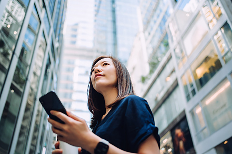 Corporate Executives Low angle portrait of confidence and successful young Asian businesswoman with coffee to go, using smartphone while standing against highrise corporate buildings in financial district in the city