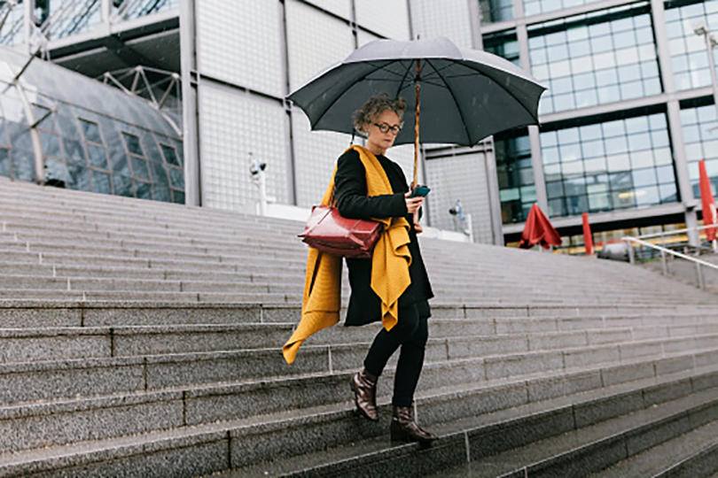 Founders A mature businesswoman walking in the rain, holding an umbrella while looking at her smartphone.