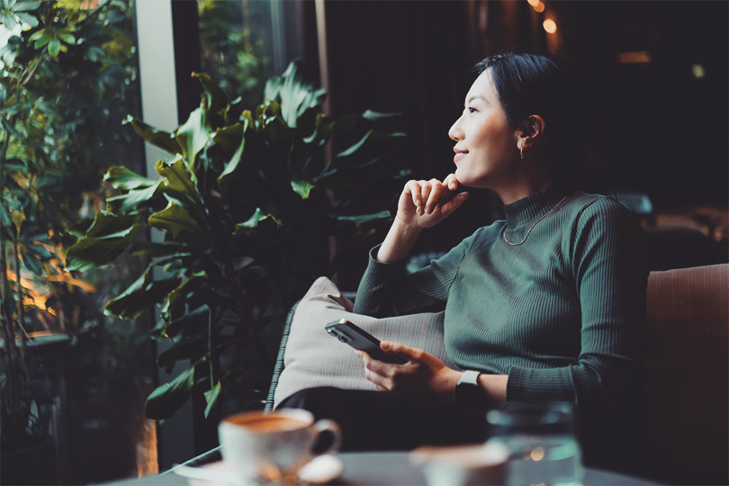  GettyImages-2149615639_Confident young Asian woman with smartphone looking out through window while sitting in a cafe having coffee. Making a personal financial plans and investment decisions. Wealth management. Business, banking, finance and investment concept