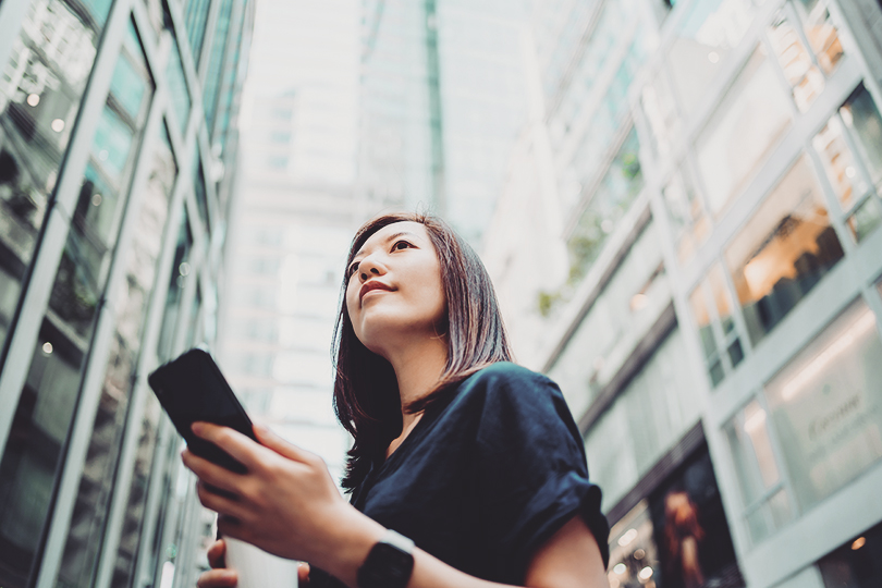 Low angle portrait of confidence and successful young Asian businesswoman with coffee to go, using smartphone while standing against highrise corporate buildings in financial district in the city