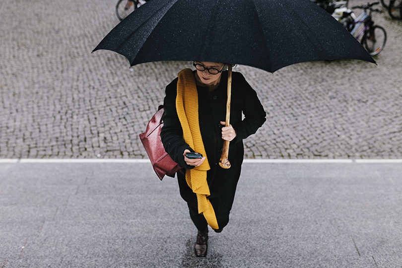 A mature businesswoman walking in the rain and holding an umbrella to protect her smartphone.