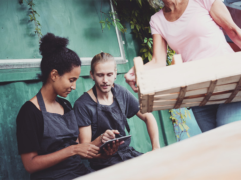 Male and female gardener using tablet computer while senior woman carrying crate in yard