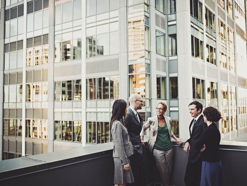 Group of coworkers standing in discussion on deck of office buildings in background