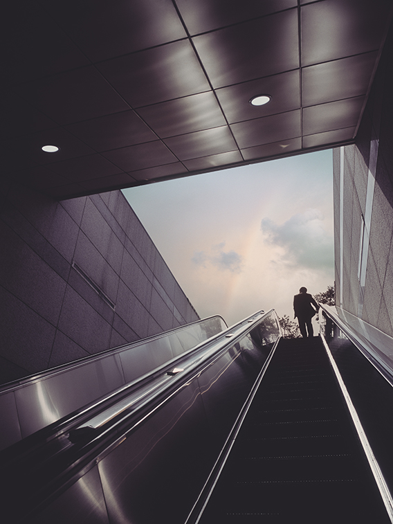 Escalator of subway station with businessman moving towards dramatic sky with rainbow.